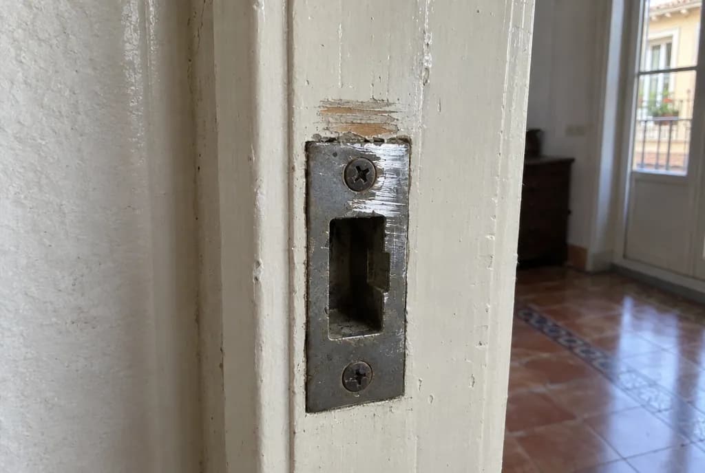 Cerradero metálico atornillado a un marco de madera pintado, con marcas de roce y pintura levantada justo encima del hueco