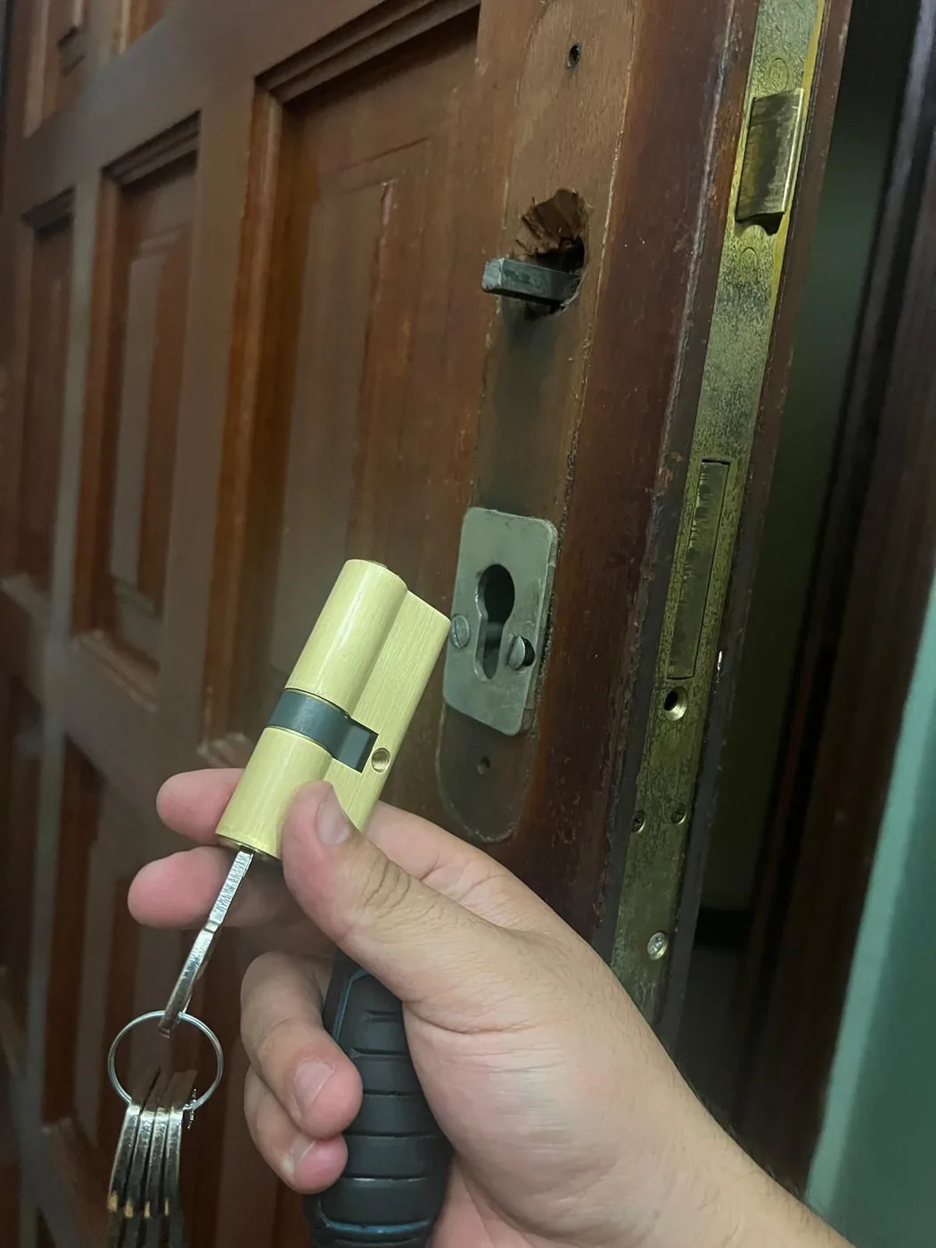 Cerrajero sosteniendo un bombín de perfil europeo nuevo frente a la cerradura embutida de una puerta de madera abierta