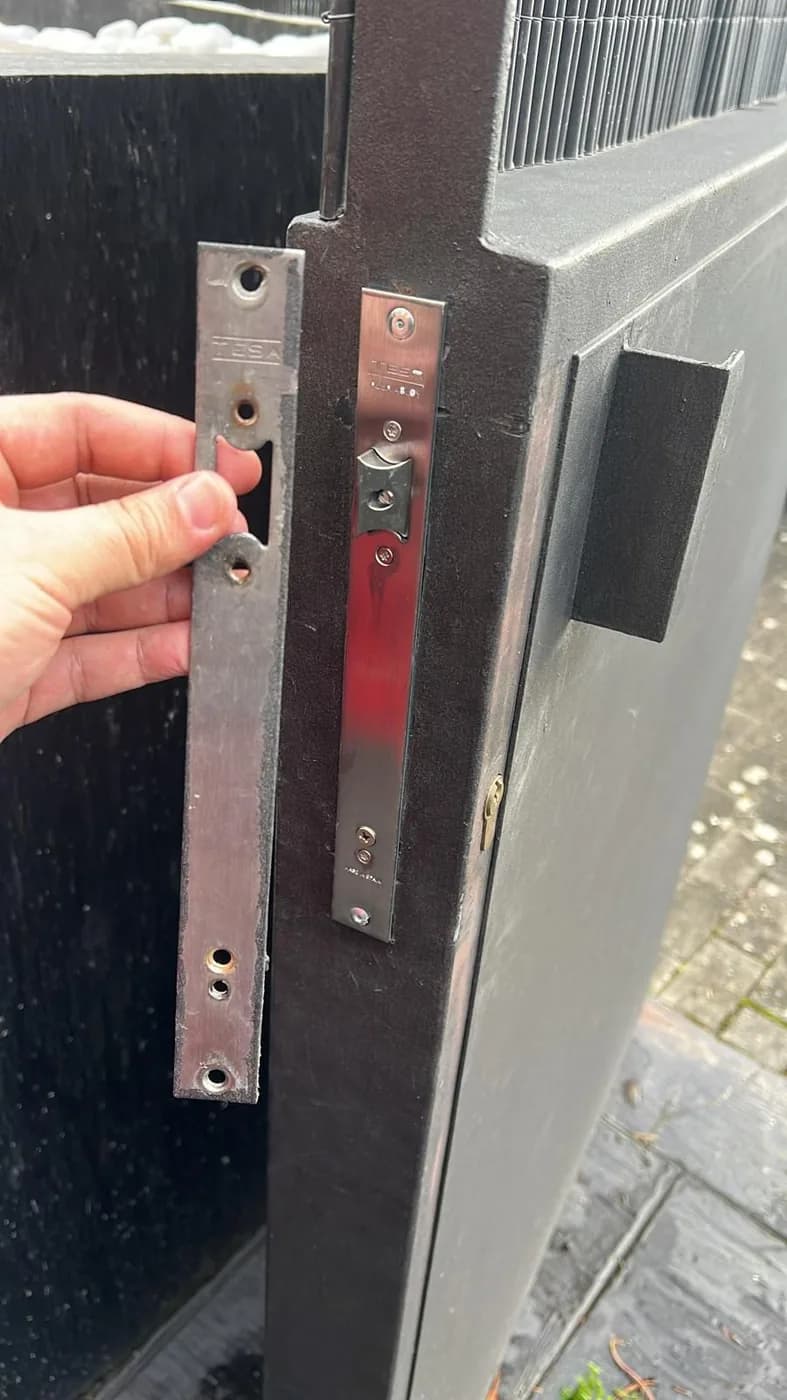 Cerradero metálico nuevo instalado en el marco de una puerta metálica exterior, con el cerradero antiguo sostenido en la mano